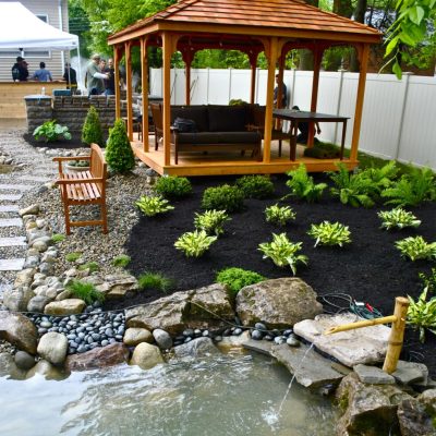 A wooden gazebo with seating overlooks a landscaped garden and small pond with rocks and flowing water.