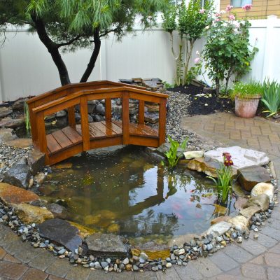 Small wooden bridge over a backyard pond with rocks, plants, and a tree, surrounded by paved paths and a white fence.