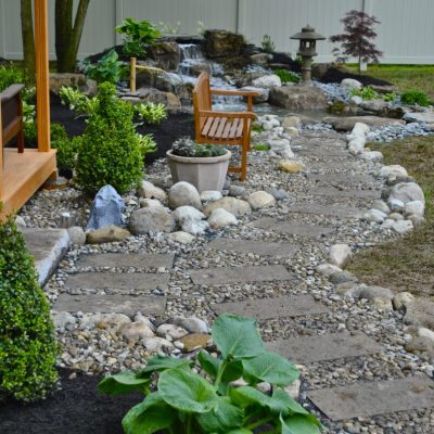 Stone pathway with bench, plants, and small waterfall in a landscaped backyard garden with a white fence.