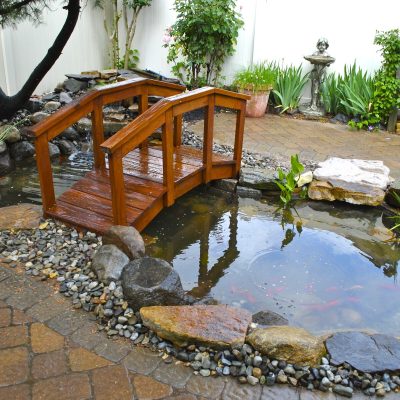 Small wooden bridge over a backyard pond surrounded by rocks, plants, and a paved patio area.