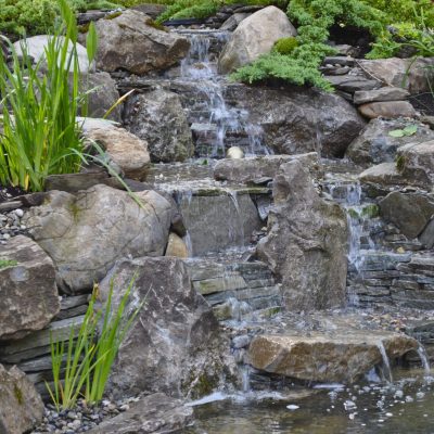 A small, tiered waterfall flows over rocks into a pond, surrounded by greenery and garden plants.