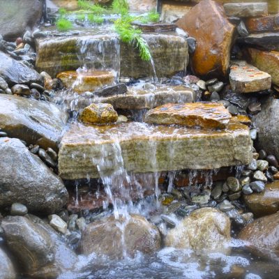 Water cascades over flat stone steps surrounded by large rocks, with green plants at the top of the falls.