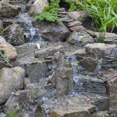 Small man-made waterfall cascading over rocks into a pond surrounded by plants and greenery in a landscaped garden.