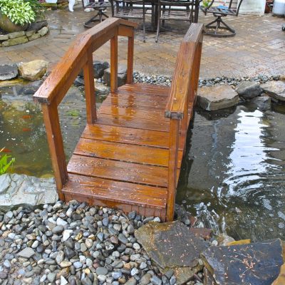 A small wooden bridge crosses over a backyard pond with rocks, plants, and several orange fish visible in the water.