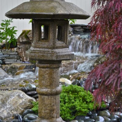 Stone lantern stands among black pebbles and greenery near a pond with a waterfall in a landscaped garden.
