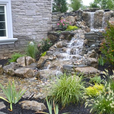 Stone waterfall feature with cascading water, surrounded by plants and flowers next to a house with a stone wall.