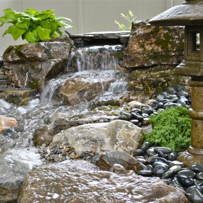 A small waterfall flows over rocks in a landscaped garden with a stone lantern and green plants.
