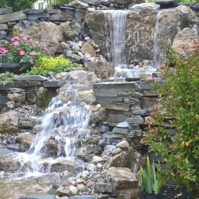 Multi-level stone waterfall feature with flowing water, surrounded by flowers and greenery in a landscaped garden.