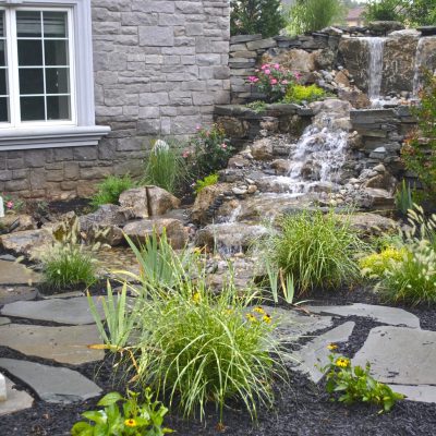 Stone patio with ornamental grasses and flowers beside a house, featuring a small waterfall and rock landscaping.