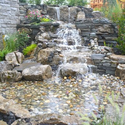 Water flows down a tiered stone waterfall into a shallow pond surrounded by rocks and garden plants.