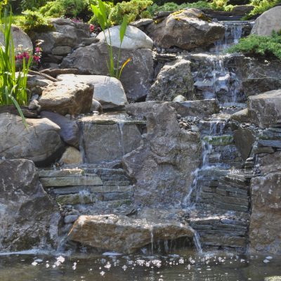 A small waterfall flows over tiered rocks surrounded by green plants and pink flowers in a landscaped garden.