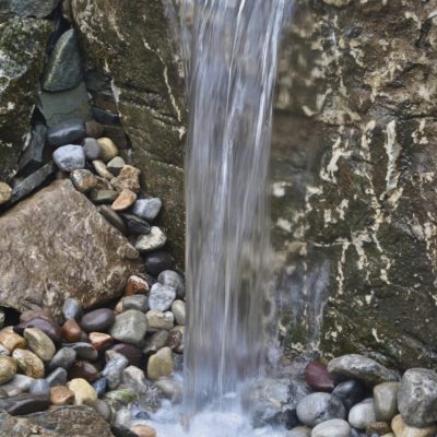 A small waterfall flows over a rock ledge onto smooth stones and pebbles below.