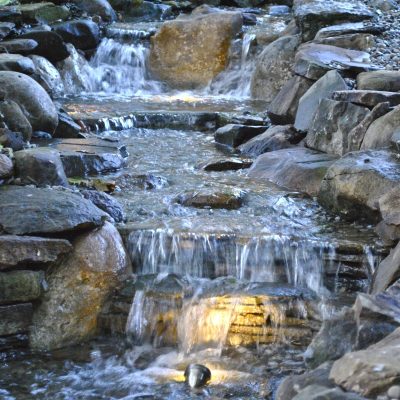 Small artificial stream with cascading water and rocks, illuminated by soft yellow lights in a landscaped garden.