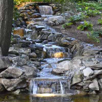 A small, tiered waterfall flows over rocks between trees, surrounded by greenery, with lights illuminating the water.