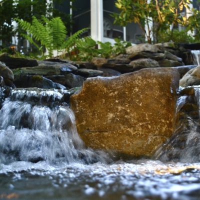 A small waterfall flows over rocks, surrounded by greenery and plants in a landscaped garden setting.