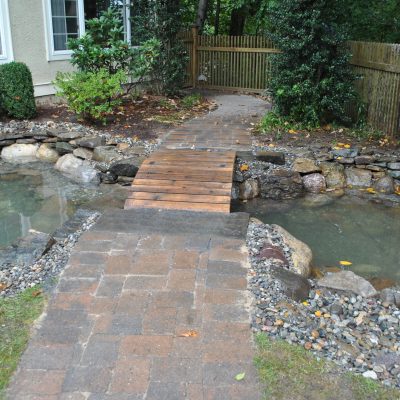 A brick and wooden footbridge crosses a small backyard pond with stone edging near a house and wooden fence.