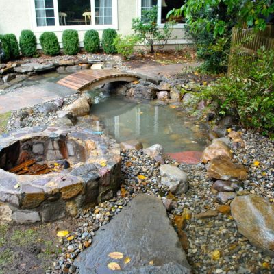 A landscaped backyard with a small pond, stone fire pit, and footbridge near a house with large windows.