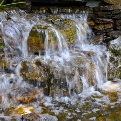 Water cascades over stacked rocks in a small man-made waterfall surrounded by green plants.