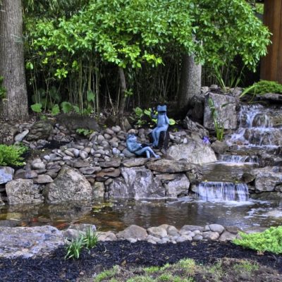 A small backyard pond with a stone waterfall, surrounded by plants, a stone bench, and a wooden building.