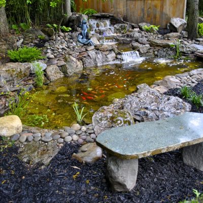 Stone bench facing a small koi pond with a waterfall, surrounded by rocks, plants, and a wooden fence in a garden.
