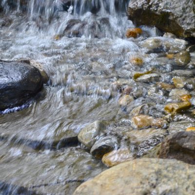 Clear water flows over smooth rocks and pebbles in a shallow stream with a small waterfall.