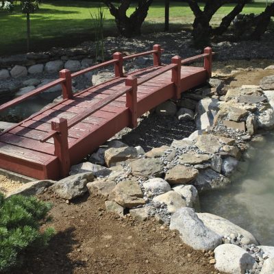 A small red wooden bridge crosses over a pond surrounded by rocks and landscaped greenery.
