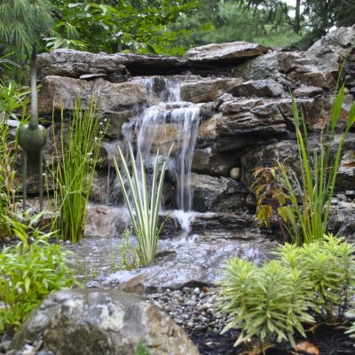 Small garden waterfall flows over rocks, surrounded by plants, a heron statue, and a gnome figurine.