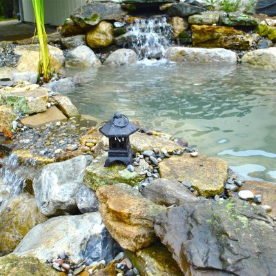 A small waterfall flows into a clear pond surrounded by rocks, with a decorative lantern placed on the edge.
