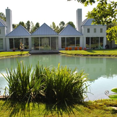 Modern house with a row of peaked roofs, large windows, and a pond with landscaping in the foreground.
