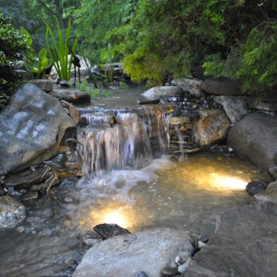 Small waterfall flows over rocks into a lit pond, surrounded by greenery in a garden setting.