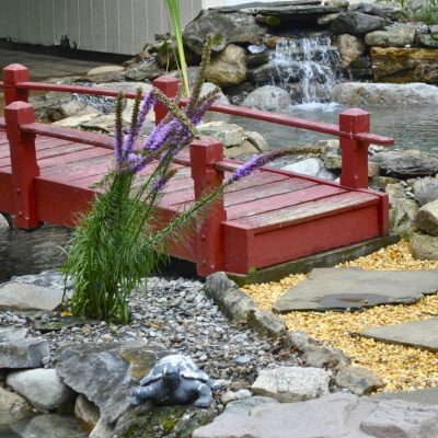 A red wooden bridge crosses over a pond with rocks, a waterfall, plants, and gravel paths in a landscaped garden.
