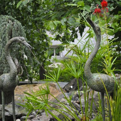 Two metal crane statues stand among green plants and rocks in an outdoor garden setting.