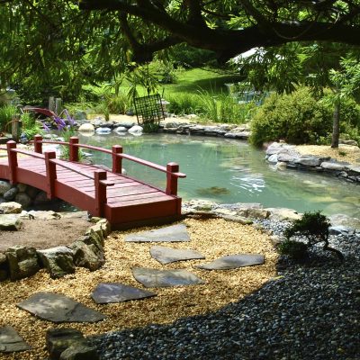 A small red bridge crosses over a pond surrounded by rocks, plants, and a gravel path in a garden.