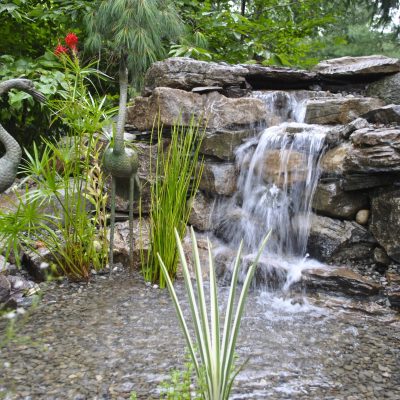 A small waterfall flows over rocks into a pond, surrounded by plants and two metal crane sculptures.