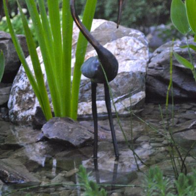 Metal bird sculpture stands in a garden pond surrounded by rocks and tall green plants.