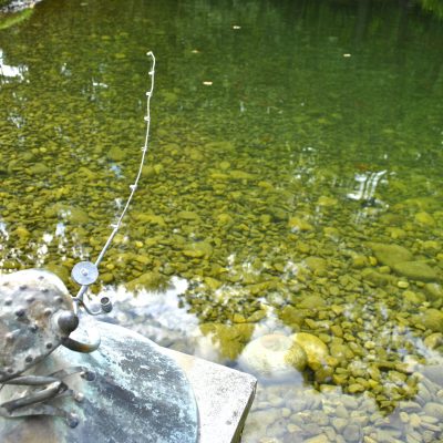 A metal frog statue holding a fishing rod sits by the edge of a clear, rocky pond.