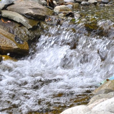 Water flows over rocks in a small, man-made stream bordered by stones and grass.