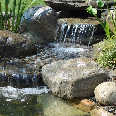 Small waterfall flowing over rocks into a pond, surrounded by green plants and pebbles in a landscaped garden.