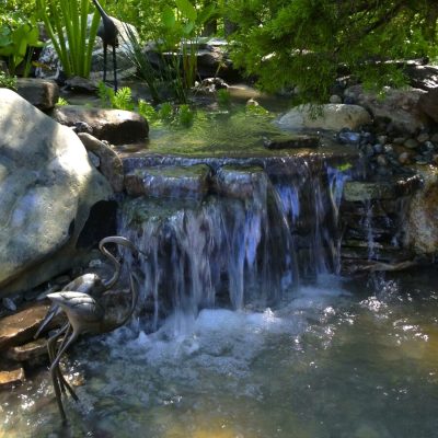 Small garden waterfall flows over rocks into a pond, surrounded by plants and metal heron sculptures.