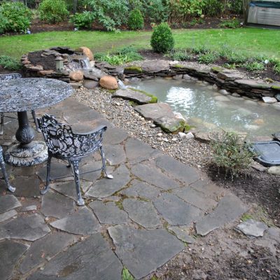 Stone patio with ornate metal table and chairs next to a small garden pond surrounded by rocks and plants.
