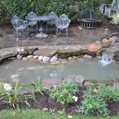 A small backyard pond with waterfalls, surrounded by stones, plants, and wrought iron patio furniture in the background.
