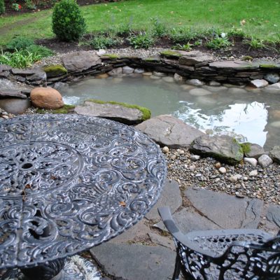 A metal patio table and chair sit near a small stone pond with water and rocks in a landscaped garden.