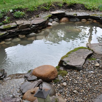 Small backyard pond surrounded by rocks, stones, and moss with water gently flowing in, bordered by green grass.