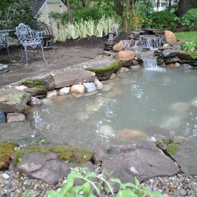Small backyard pond with rocks and a waterfall feature, surrounded by greenery and metal patio furniture on the side.