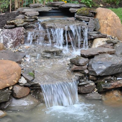 A small artificial waterfall flows over stacked rocks into a pond, surrounded by stones and greenery.
