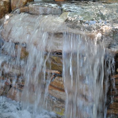 Clear water flows over stacked stones in a small waterfall feature surrounded by rocks.
