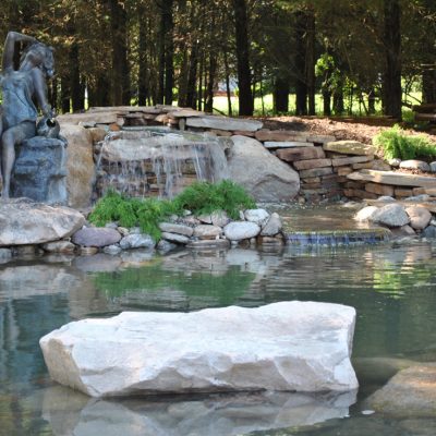 A small waterfall flows into a pond, with a statue of a woman pouring water beside rocks and greenery.