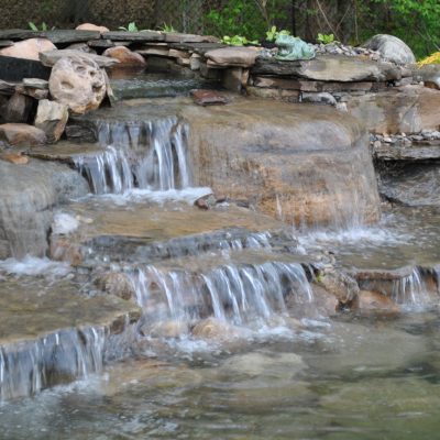 Water cascades over stone steps in a small garden waterfall surrounded by rocks and greenery.