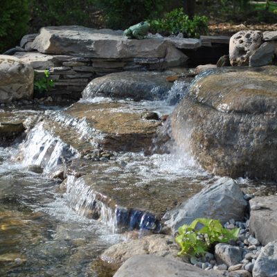 Small waterfall flows over rocks into a pond, surrounded by stones, greenery, and decorative frog statues.