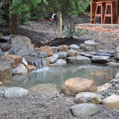A small backyard pond with rocks, a waterfall feature, and a wooden patio area with stools in the background.